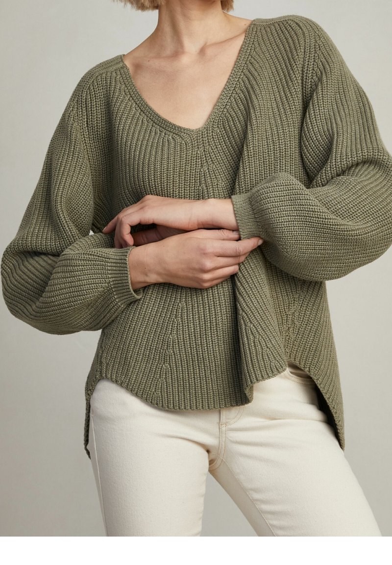 Olivgrüner Strickpullover mit tiefem V-Ausschnitt, strukturiertem Rippenmuster, Ballonärmeln und seitlichen Schlitzen. Kombiniert mit hellen Hosen.