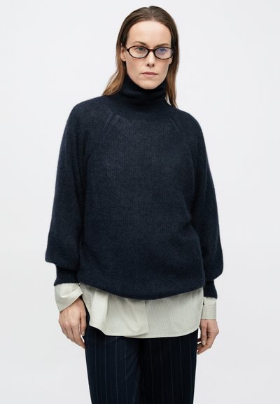 Marineblå oversized rullekravesweater lavet af blødt stof, med ribbede manchetter og en afslappet pasform, lagdelt over en lys skjorte med krave.