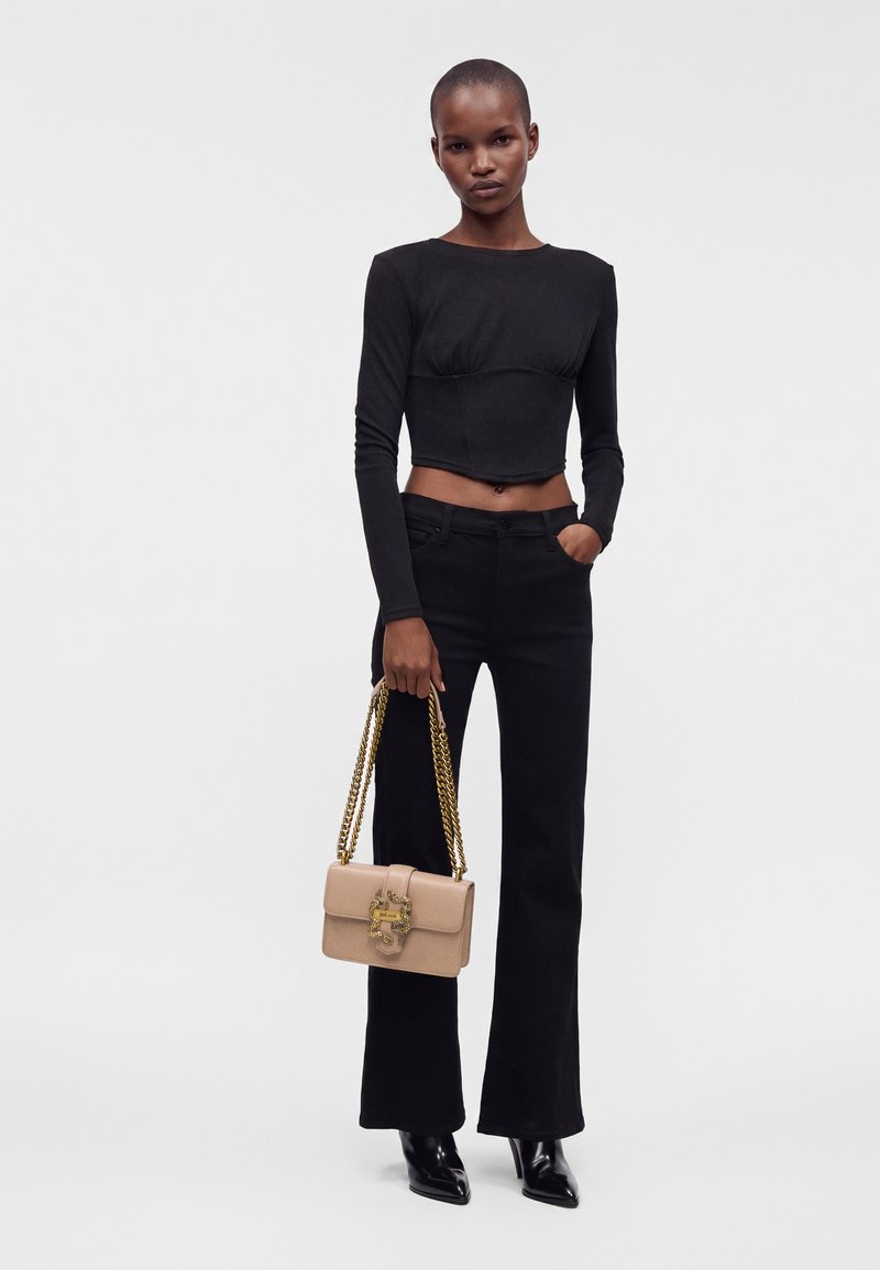 Femme mince portant un haut court à manches longues noir, un pantalon noir et des bottines noires à talons, tenant un sac à main beige avec des anses en chaîne dorée.