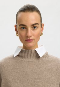 Donna con occhi azzurri e capelli raccolti all'indietro che indossa orecchini a cerchio dorati, un maglione a coste beige e una camicia bianca con colletto sotto.