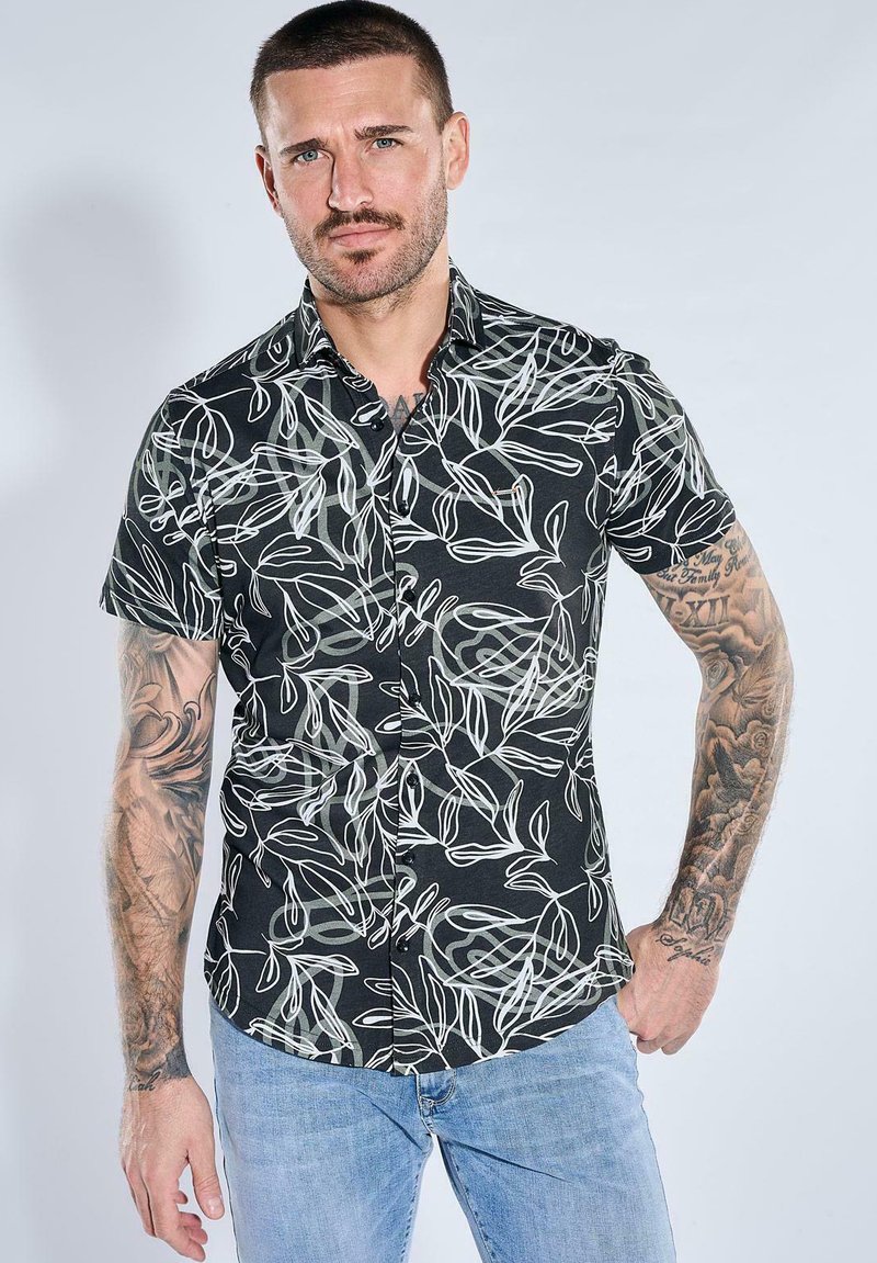Man with short hair and tattooed arms wearing a black short-sleeve shirt with white leaf patterns and light blue jeans.