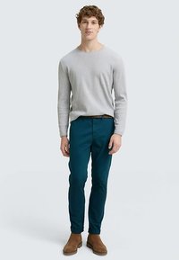 Ljusgrå ribbad tröja, teal stretchbyxor och ljusbruna mockaankelstövlar. Modellen står upprätt med händerna vid sidorna mot en neutral bakgrund.