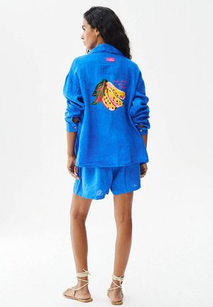 Kobieta w niebieskiej lnianej kurtce i szortach, na plecach grafika banana i tekst na kurtce, stoi na białym, jednolitym tle.