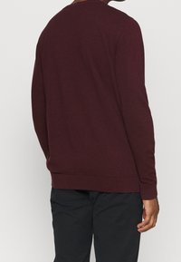 Man som bär en åtsittande, långärmad vinröd tröja och svarta byxor, sedd från ryggen mot en enkel bakgrund.