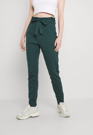 Kvinde iført mørkegrønne bukser med høj talje og bundet bælte, samt hvide sneakers, stående mod en ensartet lys baggrund.