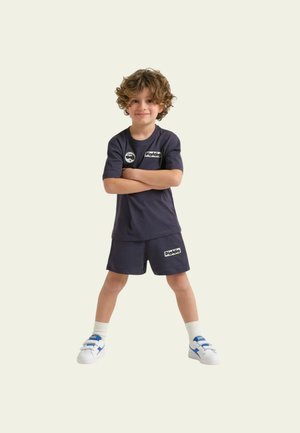 Bambino piccolo con capelli ricci in piedi con sicurezza, braccia incrociate, indossa abbigliamento sportivo blu navy e scarpe da ginnastica bianche su uno sfondo chiaro.
