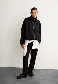 Person trägt eine schwarze Reißverschlussjacke, schwarze Hosen und Mokassin-ähnliche Schuhe, mit einem weißen Hemd um die Taille gebunden, steht drinnen.