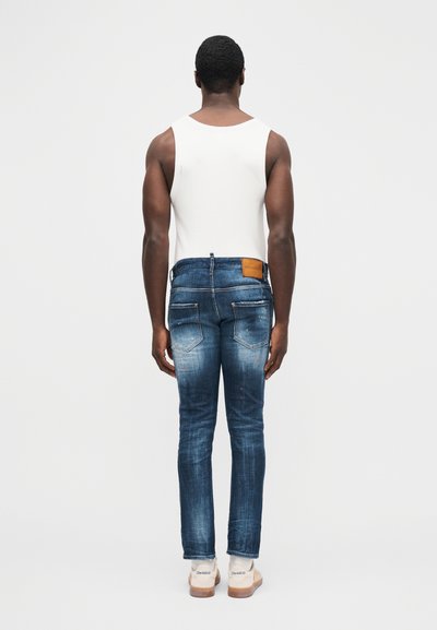 Uomo in piedi di spalle, indossa una canottiera bianca, jeans blu strappati e scarpe da ginnastica bianche su uno sfondo chiaro uniforme.