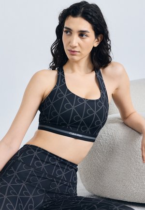 Femeie cu păr creț închis la culoare, purtând un top sport Calvin Klein negru cu model și colanți, așezată și sprijinită de un fotoliu bej texturat.