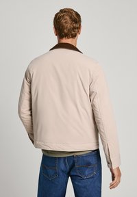 Chaqueta beige con un cuello de pana marrón, tejido suave y un diseño corto. Presenta una costura trasera sencilla y sin cierres visibles.