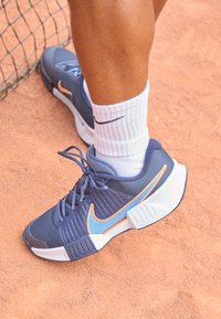 Blaue und graue Tennisschuhe aus Netzmaterial, mit strukturierter Verarbeitung, orangefarbener Nike-Welle und weißer Sohle. Getragen mit einer weißen Socke auf einem Tonplatz.