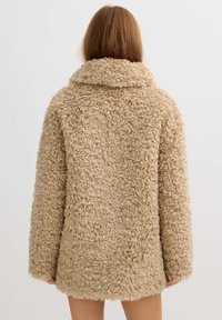 Mujer con pelo largo que lleva un abrigo de pelo sintético rizado de color beige, vista desde atrás sobre un fondo claro y liso.
