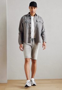 Gray button-up shirt with two chest pockets, white t-shirt, light gray shorts, and white sneakers. Model stands on wooden floor.
