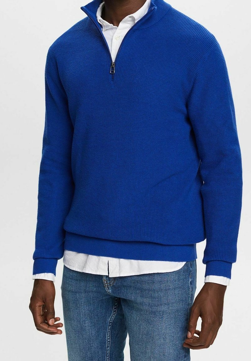 Homme portant un pull tricoté bleu roi à fermeture éclair quart sur une chemise blanche boutonnée et un jean bleu, debout devant un fond uni.