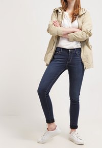 Chaqueta ligera beige sobre una blusa blanca, combinada con unos jeans skinny de color azul oscuro y zapatillas blancas. La chaqueta tiene capucha y cierre de botones.