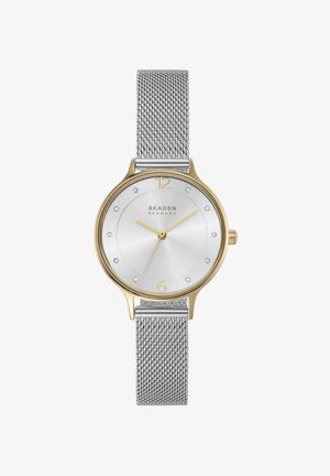 Reloj de pulsera Skagen con bisel en tono dorado, esfera plateada con efecto rayos de sol, marcadores de hora de cristal, agujas doradas y una correa de malla plateada.