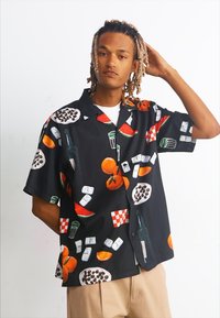 Camisa negra de manga corta con un vibrante estampado de comida y bebida, que incluye frutas, botellas y patrones a cuadros; tela ligera.