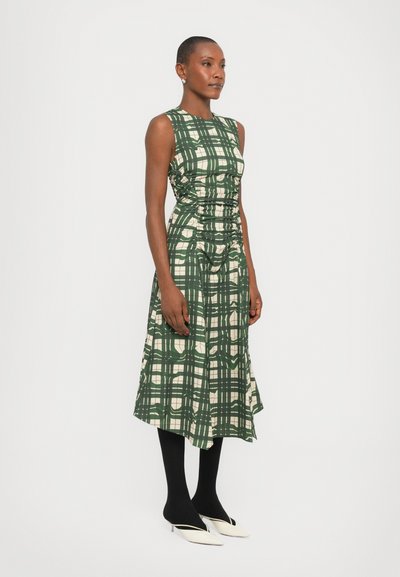 Rochie verde în carouri, cu un corset ajustat și fustă îngustată. Prezintă detalii pliate și un design fără mâneci. Poartă colanți negri și tocuri albe.