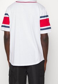 Man draagt een wit T-shirt met marineblauwe en rode horizontale strepen op de mouwen, gecombineerd met zwarte cargoshorts, gezien van achteren.