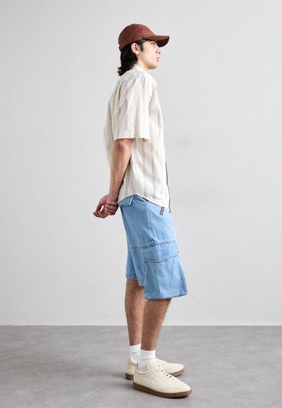Camicia a maniche corte a righe crema e blu, pantaloni cargo in denim, sneakers bianche con suola marrone chiaro e cappellino marrone indossato al contrario.