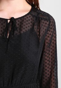 Černé šaty s texturovanými puntíky, průhlednými rukávy a zavazovacími detaily u krku. Mají našívaný pas a jemné krajkové detaily na ramenou.