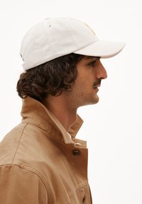 Beige pet met een gestructureerd ontwerp en een gebogen klep, gecombineerd met een bruine jas met knopen en een verhoogde kraag.