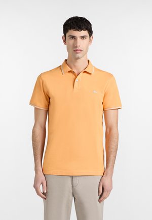 Mladić obučen u narančastu polo majicu kratkih rukava s bijelim obrubom na ovratniku i bež hlače, stoji ispred bijele pozadine.