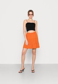 Top cropped preto com texturização em riscas e alças finas, combinado com uma saia mini em camadas vibrante na cor laranja. A modelo usa óculos de sol arredondados oversized e sapatos de salto pretos.