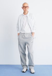 Un pantalon de survêtement gris avec une taille élastique, un t-shirt blanc et une chemise boutonnée à rayures claires. Des baskets blanches complètent la tenue.