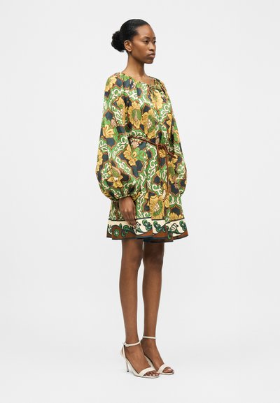 Γυναίκα στέκεται πλάγια, φορώντας φόρεμα με ζώνη και πράσινο-κίτρινο λουλουδάτο σχέδιο με φουσκωτά μακριά μανίκια, σε συνδυασμό με λευκά ψηλοτάκουνα σανδάλια.