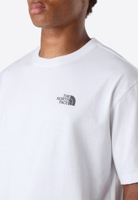 Osoba oblečená v bílém tričku s krátkým rukávem, na kterém je malé černé logo "The North Face" na levé straně hrudi, na jednolitém pozadí.