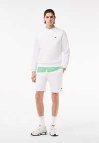 Nuori mies, joka pukeutuu valkoiseen huppariin, shortseihin, lenkkareihin ja vihreään paita, seisoo yksinkertaista valkoista taustaa vasten.