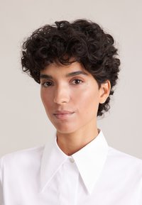 Hvid skjorte med knapåstand, stor krave, glat tekstur og en enkelt knap ved halsen. Personen har kort, krøllet, mørkt hår.