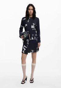 Tumesinist värvi kleit graafilise trükiga, pikad varrukad, kantud põlveni ulatuvate beežide sokkidega ja mustade sandaalidega. Kangas tundub sile, keskmise pikkusega disain.