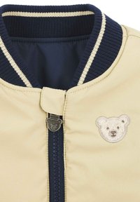 Giacca bomber beige chiaro con colletto a coste blu navy, con una toppa a forma di faccia di orso sul petto e un tirazip a forma di orso.