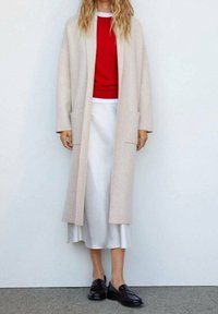 Mujer con cabello rubio largo, vestida con un abrigo largo beige sobre una blusa roja y una falda blanca, combinada con mocasines negros, de pie contra una pared lisa.