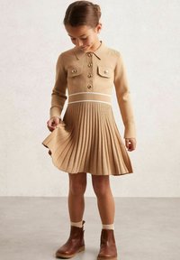 Kootud kleit kraega, täispikk plisseerkleidi seelikuosa beežis, millel on kaks rinnataskut ja pruunid nööbid. Kombineeritud pruunide pahkluudega saapade ja kreemjate sokkidega.