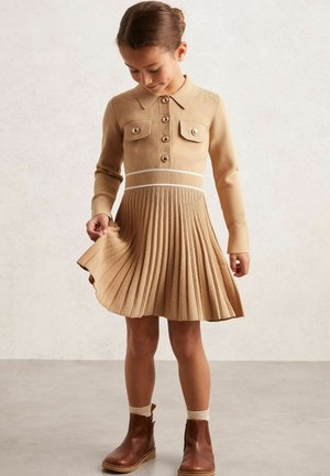 Robe en tricot avec un col, jupe entière plissée en beige, dotée de deux poches poitrine et de boutons marron. Assortie à des bottines marron et des chaussettes crème.