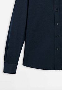 Chemise bleu marine foncé à manches longues avec manches retroussées et petits boutons noirs visibles sur la patte de boutonnage.