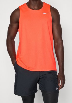 Athletische Person trägt ein helloranges ärmelloses Oberteil und schwarze Shorts mit schwarzen Leggings darunter und steht mit einer Hand in der Tasche.