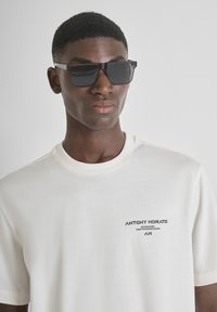 Camiseta de algodón blanca con mangas cortas, que presenta un pequeño logotipo negro en el lado izquierdo del pecho. Gafas de sol negras cuadradas con montura gruesa.