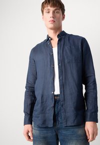 Navy blue linen shirt with a button-down collar, subtle texture, front pocket, and nude buttons, paired with light-wash denim jeans.