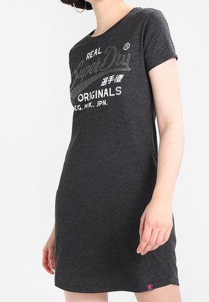 Kvinde iført en mørkegrå kortærmet kjole med teksten "Real SuperDry Originals" og japanske tegn foran mod en hvid baggrund.