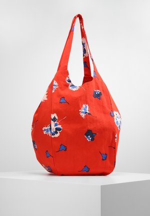 Sac fourre-tout en tissu rouge avec imprimé floral bleu et blanc, large bandoulière, présenté sur un socle blanc contre un fond uni.