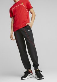 Červené tričko s bílým prošitím a logem Ferrari, kombinované s černými tepláky s červenými akcenty a černými sneakers s bílými detaily.