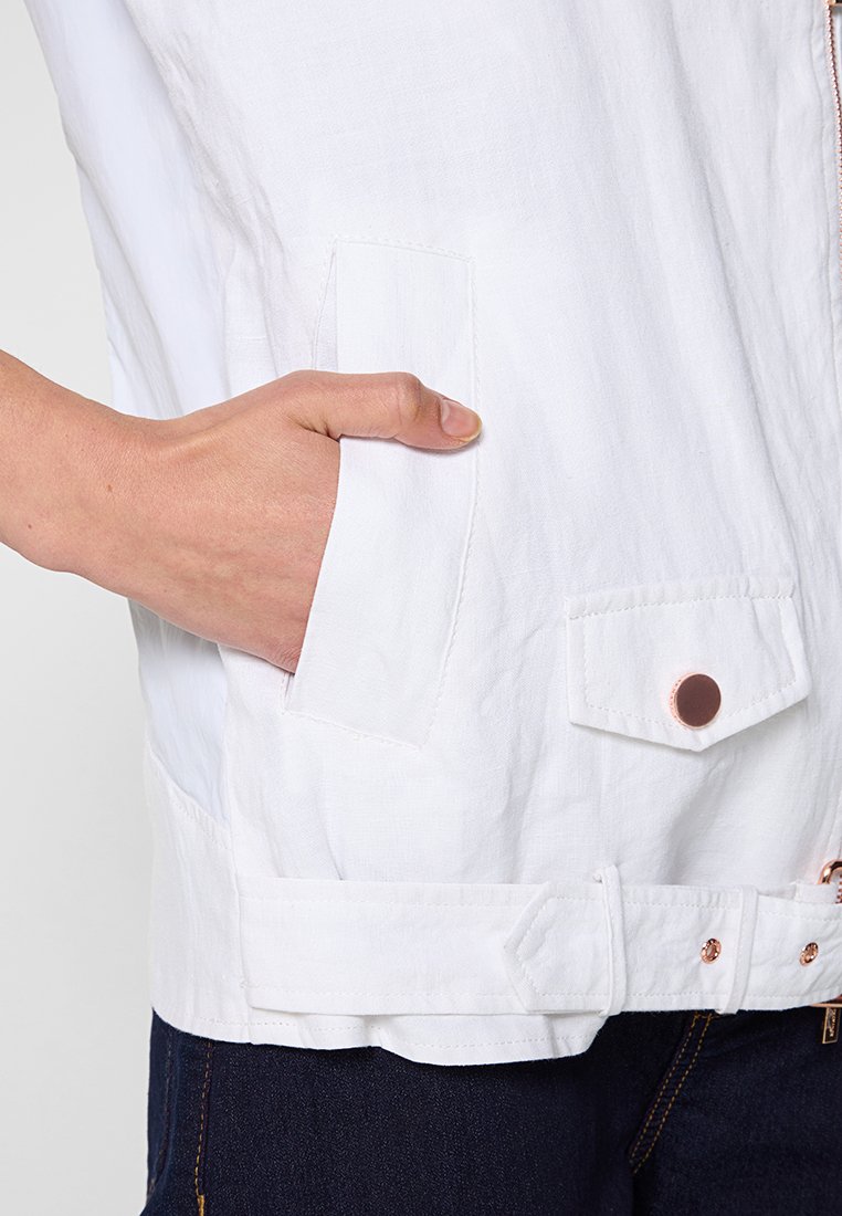 Giacca leggera bianca con zip, tasche laterali e sul petto, e dettagli in oro rosa. Presenta un design strutturato e un tessuto texture.
