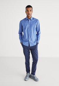 Camisa abotonada de color azul claro con un patrón sutil, combinada con pantalones azul marino y zapatillas grises. Tejido ligero, diseño con cuello.
