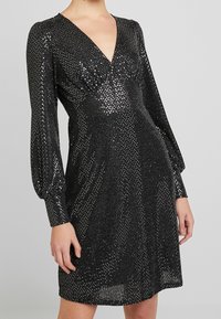 Černé šaty s hlubokým výstřihem do V, dlouhými nabíranými rukávy a lesklým stříbrným tečkovaným vzorem na strukturované tkanině.