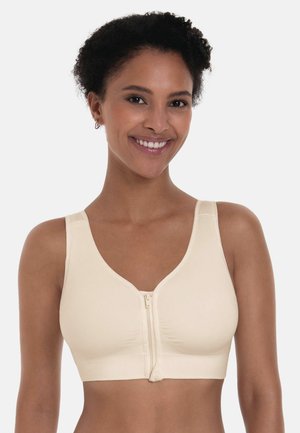 Lächende Frau trägt einen nahtlosen beige BH mit Frontreißverschluss und breiten Trägern vor einem schlichten weißen Hintergrund.