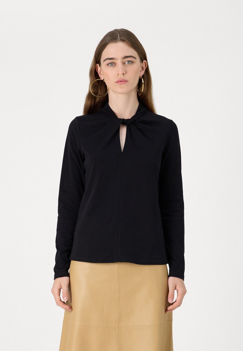 Svart langermet topp med nøkkelhulldetalj og knyttet hals. Mykt stoff, tettsittende design. Kombinert med en lys skinnskjørt.